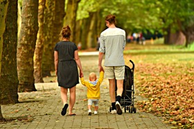 Family holding hands with a child walking along a footpath through a park with fallen leaves on the ground.