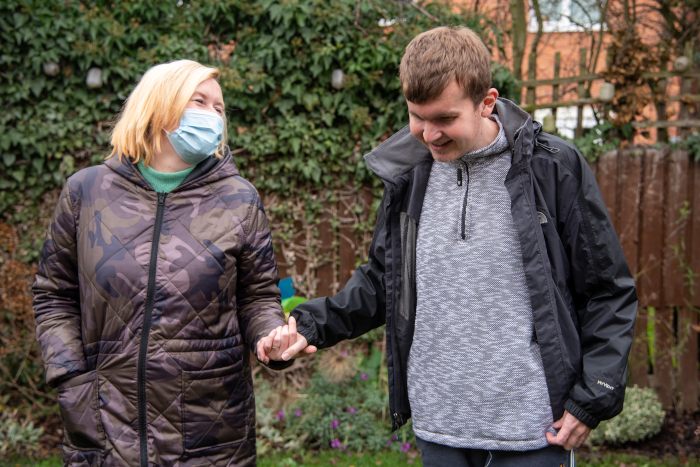 Support worker in PPE walking beside and holding hands with a young man, both smiling