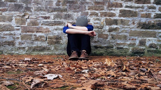 Person sitting against a wall with their head resting on their raised knees