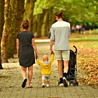 Family holding hands with a child walking along a footpath through a park with fallen leaves on the ground.