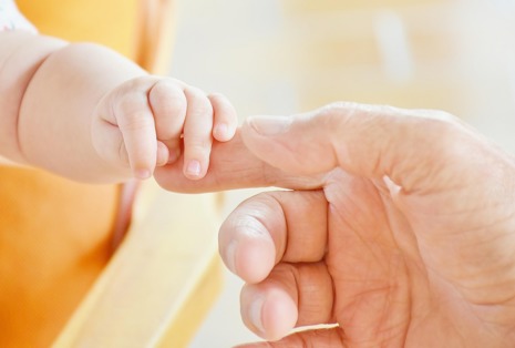 A baby holding onto an adults finger with a yellow back drop.