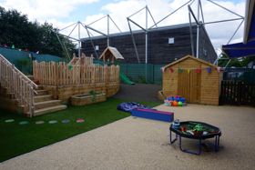 Outdoor play area with artificial grass and soft flooring, featuring a wooden climbing frame with steps, a shed decorated with bunting, a small table with play items and a stadium visible in the background.