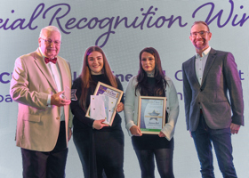 Four individuals standing on a stage during an award ceremony. Two people in the centre are holding framed certificates, while the person on the left is holding a glass trophy. The backdrop features large, partially visible text reading “Special Recognition Winners” in elegant script. All individuals are dressed in formal or semi-formal attire.