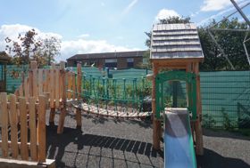 Children's outdoor play area featuring a wooden bridge structure with rope netting, a slide and safety surfacing enclosed by a green fence.