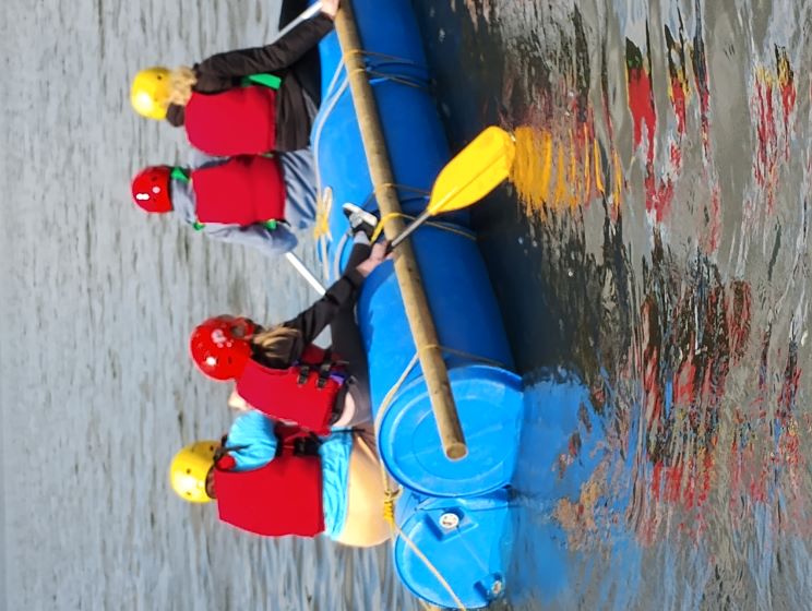 Four people in life jackets on a raft made from barrels, floating on the water.