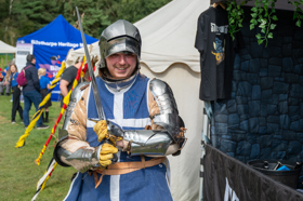 Person dressed in armour and a blue tabard holding a sword and smiling at the camera