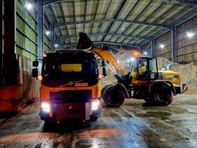 A gritting lorry at the depot being loaded with salt by a truck. There is a huge pile of salt in the barn int he background